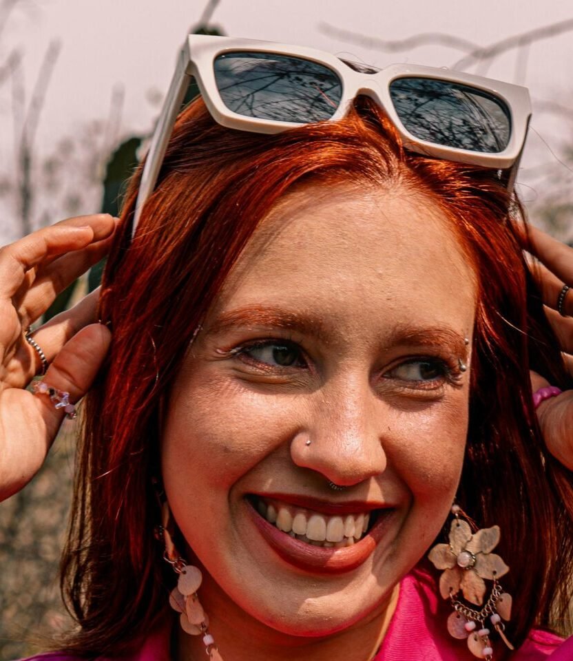 Smiling woman in a vibrant pink jacket enjoying a sunny day outdoors with stylish accessories.